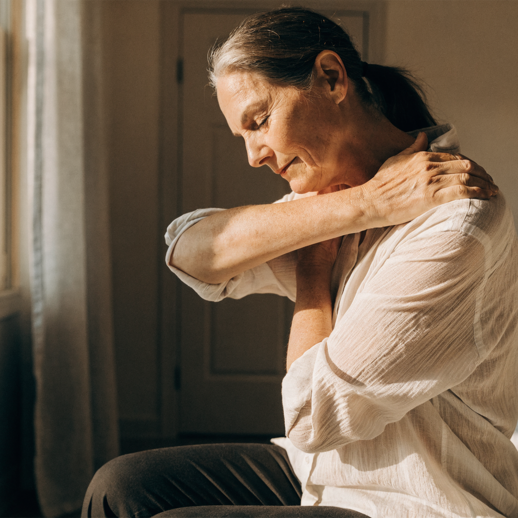 A woman sitting on the edge of a bed in morning light, doing a gentle seated shoulder stretch